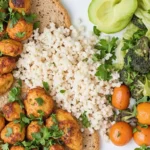 A plate of high-protein, low-carb food featuring grilled spiced chicken, cauliflower rice, whole-grain bread, and a fresh green salad with cherry tomatoes and avocado